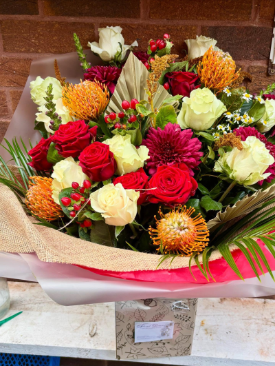 Autumn Ember Bouquet - A bold, fiery arrangement featuring rich red and creamy white roses paired with striking orange pincushion proteas and deep burgundy blooms. Accents of berry clusters, textured foliage, and golden palm leaves add depth and vibrancy.